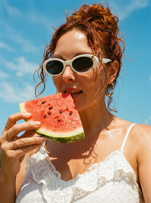 Фотореалистичный летний fashion-портрет, плёночная редакторская эстетика. Крупный план в 3/4, кадр по плечи и верх груди, камера чуть снизу, лицо в верхней части кадра на фоне неба, вертикальная ориентация. Женщина подносит крупный ярко-красный ломоть арбуза, который держит одной рукой у губ; губы слегка приоткрыты, голова чуть приподнята, взгляд в камеру, выражение чувственное, лёгкое летнее настроение. Образ: белый кружевной топ с рюшами на тонких бретелях, золотые серьги с цветными бусинами, тонкие золотые кольца. Кожа загорелая, сияющая, с лёгким влажным блеском на солнце; макияж естественный: ровный светящийся тон, выразительные брови, мягкие тени, глянцевые губы. Фон: ясное ярко-голубое летнее небо с лёгкими белыми облаками. Свет яркий прямой полуденный солнечный, тёплый, создаёт глянцевые блики на коже и сочные тени, ощущение знойного дня. Тёплая плёночная цветокоррекция, слегка винтажная и приглушённая, мелкое плёночное зерно, насыщенные тёплые тона, контраст голубого неба и тёплой кожи, мягкий контраст, глянцевый летний редакторский вайб.

Какая будет прическа?: Волосы убраны наверх в небрежный пучок, у лица свободно вьются лёгкие кудрявые пряди, слегка влажные. 

Нужны ли на фото очки? : На глазах белые ретро-очки овальной формы