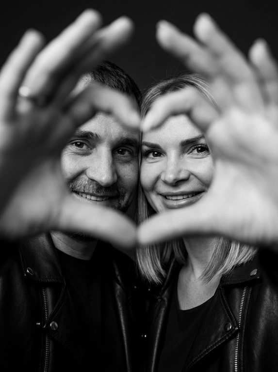 Ultra-realistic editorial photography of a smiling couple in a romantic and cinematic composition. The couple is centered in the background of the image, looking directly at the camera with a spontaneous, happy, and natural expression. In the foreground, extremely close to the lens and slightly out of focus, the couple’s hands form a heart shape perfectly framing their faces.

The composition creates strong visual depth, with sharp focus on the eyes and faces inside the heart formed by the hands. The hands occupy a large portion of the foreground, artistically blurred around the edges, creating a sophisticated cinematic effect.

Intimate, emotional, and elegant aesthetic inspired by modern perfume campaigns and premium lifestyle editorials. Soft studio lighting with delicate contrast and natural shadows. Minimalistic dark blurred background.

The couple wears minimalistic black leather jackets and sophisticated dark clothing. Genuine expressions, strong emotional connection, contemporary romantic atmosphere. Hyper-realistic skin texture, visible pores, detailed hair strands, authentic human appearance.

Black and white photography, rich tones, deep blacks, and soft highlights. 85mm lens, shallow depth of field, cinematic portrait photography, luxury editorial style, ultra detailed, hyper realistic, 8K.

Доп. характеристики: Сохранить все черты лица.У девушки волосы прямые распущены до плеч