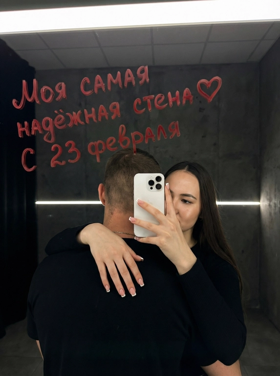 Ультрареалистичное кинематографичное зеркальное селфи пары. Строго сохранить лица из референса без изменений: возраст, пропорции, форму глаз, носа и губ, текстуру кожи, линию подбородка. Не изменять черты лица.

Локация: Серый лофт, low-key атмосфера.

Композиция: мужчина стоит спиной к камере, его лицо полностью не видно (back fully facing camera); за его спиной женщина обнимает его и снимает зеркальное селфи. Она держит белый смартфон Apple iPhone 16 Pro двумя руками. Фокус на руках и телефоне, френч квадратный маникюр. Физически корректное зеркальное отражение, без искажений..

Образ девушки: безупречный макияж, лёгкое сияние кожи.

Свет и стиль: подсветка от зеркала сверху-спереди, мягкие тени, тонкая горизонтальная световая линия на фоне. 50-85 мм, f/2-2.8, малая ГРИП; киноконтраст, плёночное зерно, high-end editorial, 8K.

Сцена полностью показана через отражение в зеркале, единственный источник изображения  — это отражение.

Ваша надпись красной помадой на зеркале: Моя самая надёжная стена❤️ С 23 февраля

Ваша одежда: Мужчина и женщина одеты в чёрной рубашке;