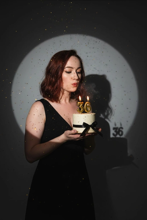 Фотореалистичный портрет женщины с точно таким же лицом, чертами, выражением и цветом волос, как на исходной фотографии - ничего не изменять. Атмосфера - день рождения: мягкий блеск свечи отражается на её лице, в воздухе лёгкие золотистые блёстки, создающие праздничное сияние. Фон темно-серый. Освещение студийное, контрастное, с кругом света на фоне, создающим выразительную тень. Волосы - волнистые, мягко подсвечены. Она немного стоит полубоком. Женщина держит в руках маленький праздничный торт, украшенным чёрными лентами и изящной цифрой из золотых свечей; Она сдувает свечу. Она одета в черное платье без рукавов. Свечки в виде цифр 36