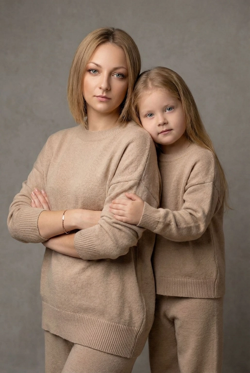 Используй эти два фото мамы и дочери и создай для них студийный портрет в чистом и элегантном стиле. Они стоят рядом, очень близко друг к другу. Дочь мягко обнимает маму за руку и слегка к ней прижимается со спины. Мама сложила руки перед грудью. Образ минималистичный, современный и стильный. На руке у матери тонкий золотой браслет. Профессиональный макияж и прическа.  Не менять черты лица ни на 1 процент. Фон — нейтральный серый с мягкой текстурой. Освещение рассеянное, мягкое, подчёркивающее естественные тона кожи. Атмосфера тёплая, нежная и спокойная, в стиле журнальной съёмки. Обе в свободных стильных бежевых свитерах и брюках.