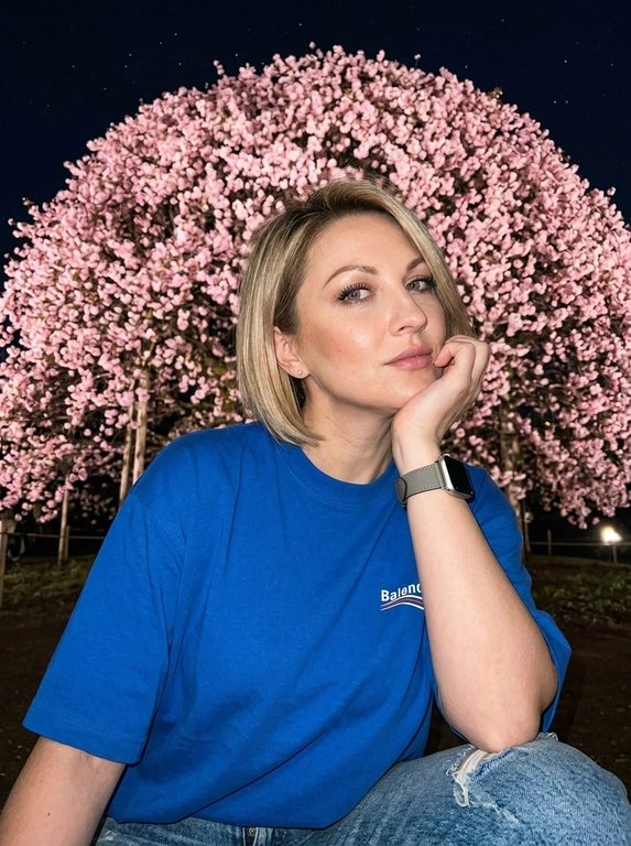 На снимке запечатлена женщина в задумчивой и мечтательной позе ночью, она сидит вполоборота в профиль, мягко подпирая подбородок рукой и устремляя нежный взгляд в камеру, лёгкая динамика ветра, subtle motion blur, темнота, композиция выстроена с драматичного слегка нижнего ракурса, где доминирующим элементом фона выступает пышная крона цветущей сакуры в ночное время, множество нежно-розовых соцветий образуют над головой плотный живой купол, подсвеченный мягким искусственным светом, создавая сказочную интимную уединённую атмосферу ночи, женщина одета в свободную футболку Balenciaga, внизу джинсы свободного кроя из плотного денима с потертостями, на руке часы Apple Watch с ремешком Hermes серого цвета, лицо освещено жёсткой вспышкой которая с гиперреалистичной точностью передает каждую деталь: естественный микрорельеф кожи, изгиб длинных ресниц и нежный макияж с акцентом на губах, палитра объединяет глубокий чёрный цвет ночного неба со звездами и пудрово-розовые тона лепестков, все микродетали кожи и текстуры материалов проработаны с высокой чёткостью, фотореализм, waist shot, low angle, 35mm, iPhone 16 Pro Titanium, гиперреалистичная кожа с видимыми порами и естественными несовершенствами.

Какого цвета будет футболка?: На ней надета синяя футболка