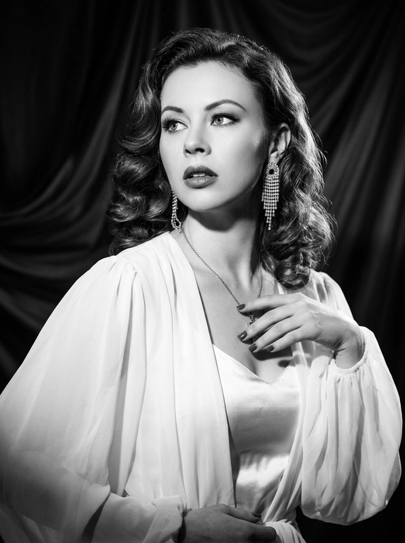 Фотореалистичный контрастный чёрно-белый glamour/editorial портрет в стиле Old Hollywood, вертикальный кадр по грудь. Женщина в 3/4, корпус чуть наклонён вперёд; голова повернута влево, взгляд вверх/в сторону (не в камеру), губы чуть приоткрыты, выражение драматичное и кинематографичное. Образ: белая накидка из шифона с длинными струящимися рукавами, под накидкой драпированный атласный белый топ; на ушах крупные серьги со стразами/каскадом. Макияж: классический вечерний glam (в ч/б читается как контраст) — чёткие брови, мягкие smoky тени, тонкая стрелка, длинные ресницы, губы с чётким контуром и сатиновым финишем. Фон: тёмный студийный с мягким рисунком/драпировкой (как бархатные шторы), размытый. Свет: жёсткий key слева спереди (beauty dish) + контровой/rim справа для отделения волос, глубокие тени, яркие блики на скулах и волосах, богатый тональный диапазон (глубокие чёрные и сливочные белые). Камера: 85mm, f/2.0–2.8, ISO 100–200, 1/160, резкость на глазах и губах, фон в мягком боке, лёгкое плёночное зерно, без “пластика”.

Какая будет укладка? : Волосы: объёмные голливудские волны, аккуратная укладка с сильным объёмом и мягкими завитками, блеск волос.