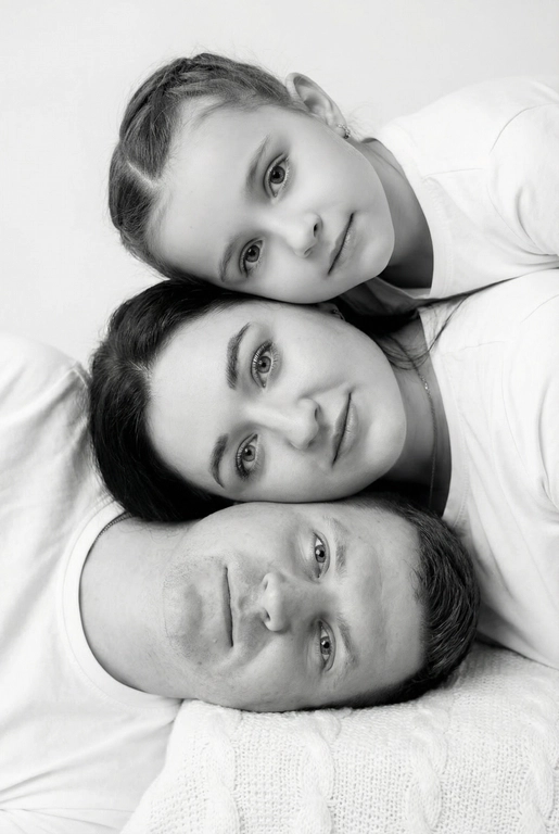 На основе предложенных фотографий, создай профессиональный студийный портрет для этих людей, снятый сверху (top-down, overhead shot). Сохрани настоящие лица, возраст и индивидуальные черты людей.
Композиция «снизу вверх», шахматный порядок:
﻿﻿самый старший человек находится внизу, как мягкая подушка, его голова опирается на нейтральную студийную поверхность (например, плед или подушка);
следующие люди аккуратно и легко расположены на старшем человеке в шахматном порядке, их голова слегка приподнята, без ощущения давления; ﻿﻿

Несмотря на шахматное расположение, головы выстроены строго в одну вертикальную линию, взгляд в камеру.
Расположи их по убыванию возраста.

Видны только головы и плечи. Выражения мягкие, спокойные, нежные. Одежда однотонная, светлая или нейтральная, элегантная, без чёрного цвета.
Фон - чистый студийный, без пола и лишних деталей, создающий эффект «воздушного» портрета.
Стиль и освещение:
Мягкий ровный свет сверху, минимум теней. Чистый фон; фотореализм, высокая детализация лиц, аккуратная текстура кожи; 8K.
 Студийная черно-белое фотография. На ребенке белая рубашка. На мужчине белая рубашка. На женщине белая рубашка