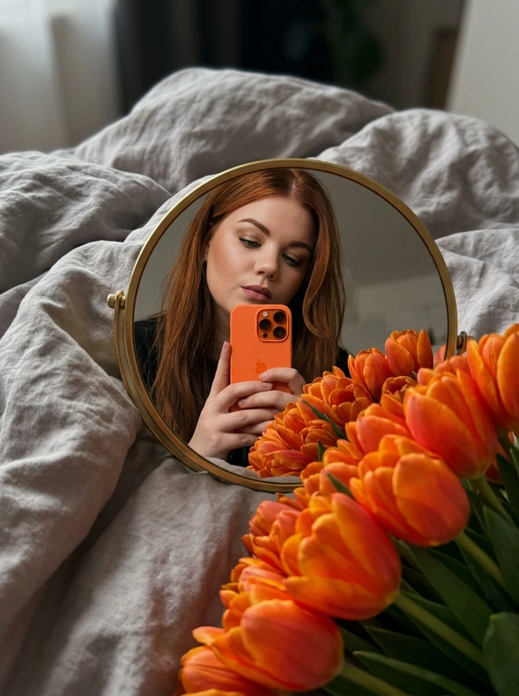 Создай портрет, не меняя черты лица. Фотография в стиле селфи в зеркале, отражающая женщину с безупречным макияжем. На переднем плане — огромный, роскошный букет цветов, которые занимают значительную часть кадра, Женщина держит оранжевый iPhone 17 PRO. Зеркало компактное круглой формы в золотой оправе лежит на белой кровати, женщина склоняется над ним, ракурс снизу. 
Атмосфера и детали:
Фон — мягкое серое постельное белье с естественными складками, создающее уютную домашнюю обстановку. Освещение мягкое, приглушенное, с акцентом на насыщенный цвет цветов. Композиция кадра динамичная: зеркало расположено под углом, создавая интересную перспективу и глубину.
Технические параметры:
Высокая детализация, реализм, текстура лепестков цветов и ткани, глубина резкости (bokeh) на заднем плане, эстетика Pinterest, 8k, кинематографичное качество.

Букет: Роскошные оранжевые тюльпаны