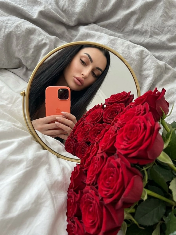 Создай портрет, не меняя черты лица. Фотография в стиле селфи в зеркале, отражающая женщину с безупречным макияжем. На переднем плане — огромный, роскошный букет цветов, которые занимают значительную часть кадра, Женщина держит оранжевый iPhone 17 PRO. Зеркало компактное круглой формы в золотой оправе лежит на белой кровати, женщина склоняется над ним, ракурс снизу. 
Атмосфера и детали:
Фон — мягкое серое постельное белье с естественными складками, создающее уютную домашнюю обстановку. Освещение мягкое, приглушенное, с акцентом на насыщенный цвет цветов. Композиция кадра динамичная: зеркало расположено под углом, создавая интересную перспективу и глубину.
Технические параметры:
Высокая детализация, реализм, текстура лепестков цветов и ткани, глубина резкости (bokeh) на заднем плане, эстетика Pinterest, 8k, кинематографичное качество.

Букет: Роскошные красные розы