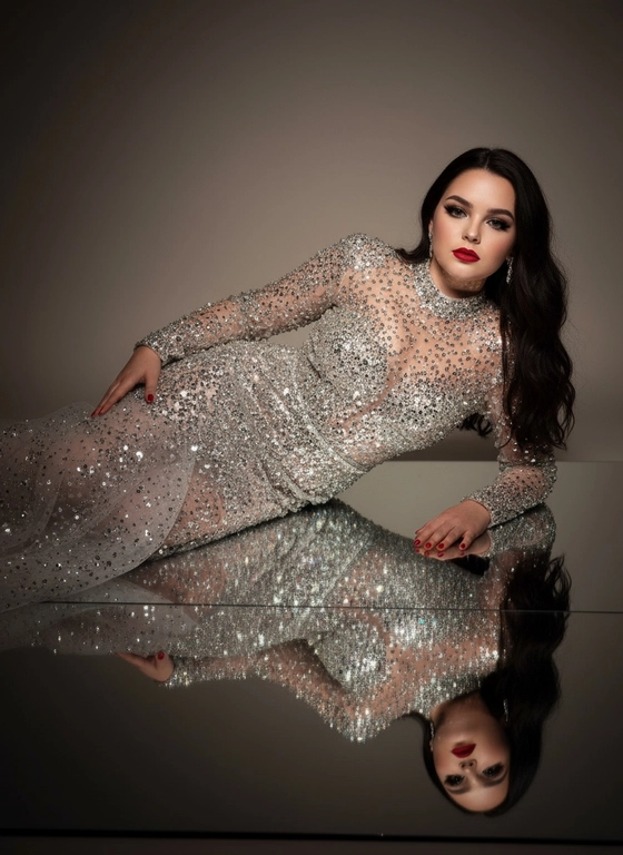 Это изысканный портрет женщины, лежащей боком на зеркальной поверхности, создавая идеальное отражение всего ее тела. На ней элегантное, облегающее платье от кутюр, полностью расшитое серебряными стразами, кристаллами и сверкающими деталями, которые роскошно ловят свет. Платье имеет длинные рукава, высокий ворот и струящийся шлейф из прозрачного тюля, усыпанного стразами на юбке, источающий гламур и изысканность. У нее длинные, волнистые волосы, которые нежно падают на ее плечи. Ее макияж безупречен с яркими темными smoky eyes и черной подводкой для глаз, длинными, объемными ресницами, безупречной кожей и пухлыми губами с изысканной, яркой красной помадой. На ней сдержанные, но элегантные серьги, дополняющие гламурный образ. Сцена драматически освещена темной виньеткой, подчеркивающей блеск платья и контраст между ее телом и отражением в зеркале. Минималистичный фон в нейтральных тонах ещё больше подчёркивает харизму модели и богатство деталей наряда. Общая атмосфера создаёт ощущение роскоши, элегантности и изысканности, отражая моду и высокую моду. Сохранены те же черты лица без каких-либо изменений, тот же тон кожи и цвет волос. Качественный маникюр, профессиональное студийное качество изображения.