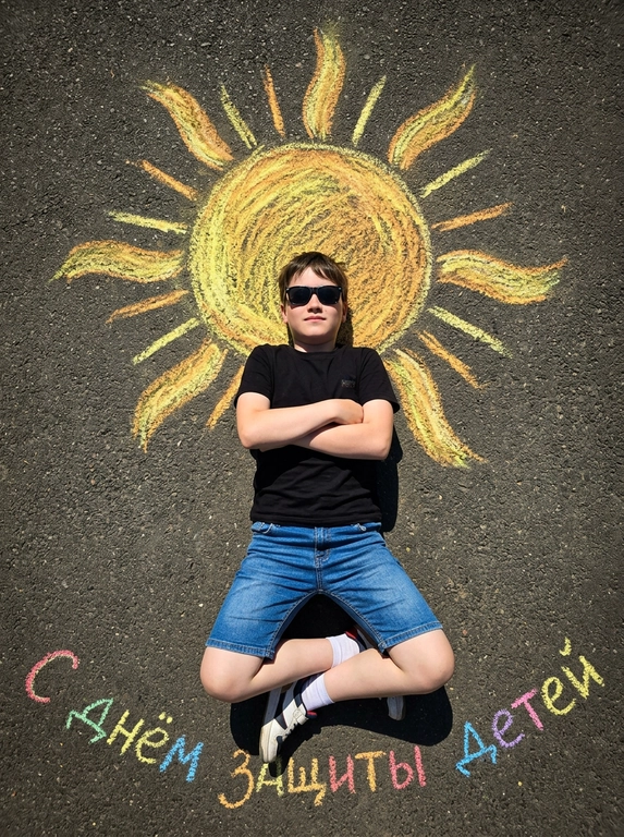 Top-down shot of a child lying on dark grainy asphalt. A bright chalk-drawn sun with radiant rays is drawn above the child’s head. The child is wearing black sunglasses, a black t-shirt, denim shorts, and sneakers with white socks. Arms crossed on chest, legs bent and crossed at ankles. Vibrant chalk sun texture in yellow, orange, and warm golden tones. Harsh midday sunlight, sharp shadows, high contrast. Summery playful atmosphere. On the asphalt below, colorful chalk lettering reads "С днем защиты детей". Photorealistic, 8k, highly detailed texture.