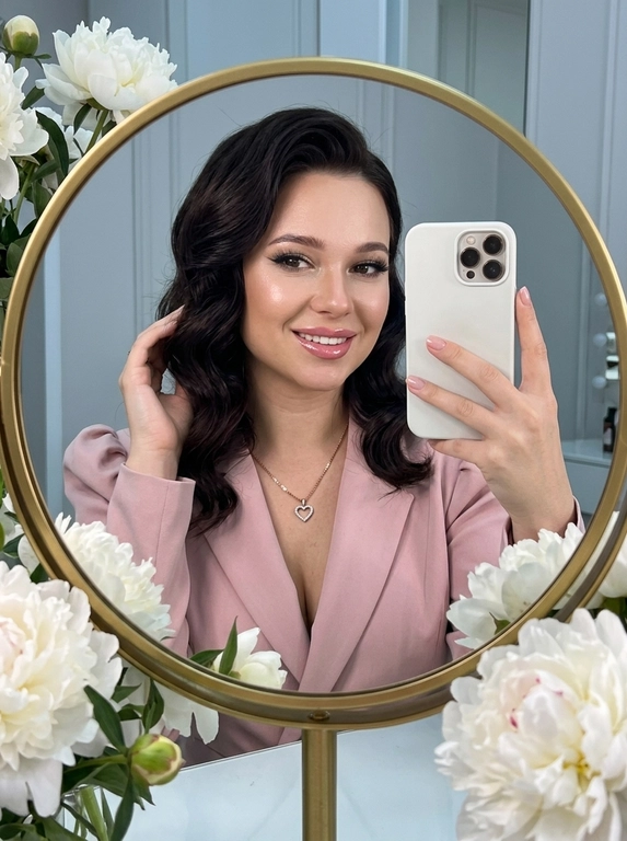 Максимально реалистичное селфи в зеркале в эстетике luxury soft glam. Модель стоит перед круглым зеркалом с изящной рамой, украшенной белыми живыми пионами. Цветы обрамляют зеркало нежно и дорого, создавая ощущение утончённого, премиального кадра. Композиция чистая и визуально собранная: модель находится в центре отражения, кадр выглядит эстетично, дорого и современно.

Поза естественная и уверенная: одной рукой модель держит современный смартфон, второй рукой слегка касается волос, создавая живой и непринуждённый жест. Селфи выглядит не постановочным, а как красивый спонтанный кадр из дорогой life-style съёмки.

Макияж — выразительный soft glam: чёткие чёрные стрелки, длинные пышные ресницы с лисьим эффектом, внутренние уголки глаз подсвечены сияющими тенями. Кожа ровная, ухоженная, с мягким влажным сиянием. Губы подчёркнуты карандашом и покрыты полупрозрачным глянцевым блеском, создающим аккуратный объём.

На модели ее образ выглядит женственно, современно и дорого. 

Смартфон современный, в минималистичном белом матовом чехле. Если нужен более дорогой вайб, без лишнего декора и ярких деталей.

Освещение холодное, мягкое и чистое, как в дорогой beauty-съёмке или premium dressing room. Свет красиво подчеркивает сияние кожи, текстуру ткани, свежесть пионов и блеск украшения. Атмосфера — роскошная, женственная, ухоженная, без дешёвого глянца.

Фотореализм максимальный: высокая детализация кожи, ткани, цветов, аксессуаров и отражения в зеркале. Лёгкий цифровой шум, мягкая резкость, деликатная зернистость, естественная глубина кадра, высокая контрастность, эффект живого фото, без CGI и без искусственного AI-эффекта.

Какая будет прическ?: крупные голливудские волны, мягкий блеск волос, аккуратная укладка на одну сторону.

Какой будет образ на фото? : Элегантный пудрово-розовый жакет с акцентными рукавами-фонариками и глубоким V-образным вырезом. На шее — тонкая изящная цепочка с ювелирным кулоном в форме сердца.