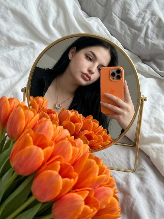 Создай портрет, не меняя черты лица. Фотография в стиле селфи в зеркале, отражающая женщину с безупречным макияжем. На переднем плане — огромный, роскошный букет цветов, которые занимают значительную часть кадра, Женщина держит оранжевый iPhone 17 PRO. Зеркало компактное круглой формы в золотой оправе лежит на белой кровати, женщина склоняется над ним, ракурс снизу. 
Атмосфера и детали:
Фон — мягкое серое постельное белье с естественными складками, создающее уютную домашнюю обстановку. Освещение мягкое, приглушенное, с акцентом на насыщенный цвет цветов. Композиция кадра динамичная: зеркало расположено под углом, создавая интересную перспективу и глубину.
Технические параметры:
Высокая детализация, реализм, текстура лепестков цветов и ткани, глубина резкости (bokeh) на заднем плане, эстетика Pinterest, 8k, кинематографичное качество.

Букет: Роскошные оранжевые тюльпаны