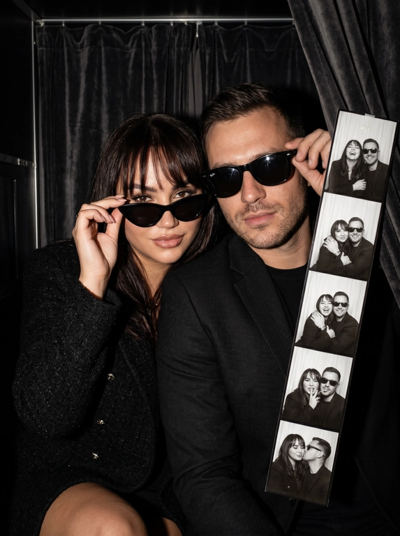 Крупный план (close-up) эффектной пары — женщины и  мужчины (не меняй лица), позирующих внутри тесной винтажной фотокабины. Они сидят вплотную друг к другу, создавая атмосферу ночного города, свидания или светской вечеринки. Композиция плотная и динамичная.

Одежда и стиль:
*   Женщина (слева): В классическом черном пиджаке. Одной рукой она кокетливо поправляет свои черные солнцезащитные очки. У нее безупречный вечерний макияж с влажным блеском на губах.
*   Мужчина (справа): Он одет в строгий черный пиджак, на нем также надеты классические черные солнцезащитные очки (Wayfarer или авиаторы), которые он слегка придерживает рукой.
*   Образ пары выдержан в едином стиле «Total Black» и эстетике «Old Money» или «Secret Agent».

Окружение и детали:
Действие происходит внутри аутентичной фотокабины. Справа на переднем плане видна свисающая длинная лента готовых черно-белых фотоснимков (photobooth film strip), на которых запечатлена эта же пара в разных позах. Фон темный, с тяжелыми складками занавески, что подчеркивает интимность момента.

Стиль и освещение:
Освещение имитирует резкую вспышку фотокабины — яркий фронтальный свет, создающий глубокие тени и подчеркивающий текстуру кожи и блеск аксессуаров. Цветовая палитра приглушенная, монохромная с теплыми телесными оттенками. Атмосфера «Nightlife Luxury».

Высочайшая детализация, акцент на блики в очках и текстуру ткани пиджаков, реалистичные черты лиц, кинематографичное качество, гиперреализм, 8k.