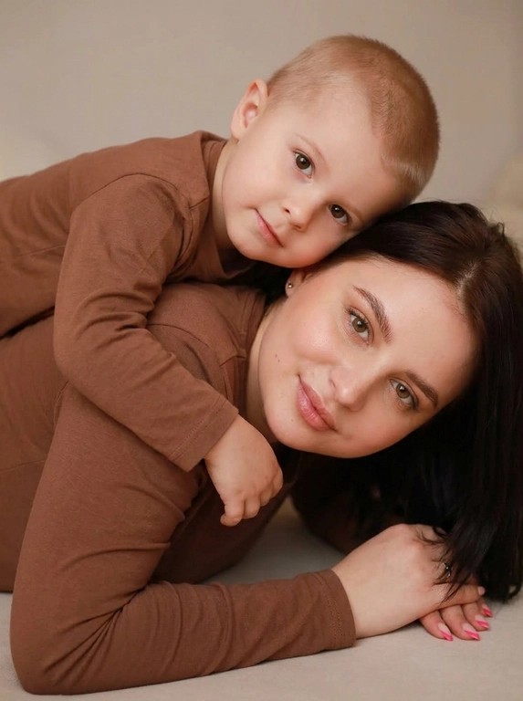 Создай студийный портрет родителя и ребёнка, максимально точно сохранив черты лица и выражения с референсных изображений. Не изменяй и не искажай лица, воспроизведи их с фотографической точностью.

Композиция:

Крупный план, ракурс на уровне глаз.

Родитель и ребёнок лежат на животе, расположены боком к камере.

Ребёнок лежит на спине родителя и мягко обнимает его за плечи.

Оба смотрят прямо в камеру, создавая тёплый и близкий зрительный контакт.

На родителе и ребёнке одинаковые коричневые лонгсливы без принтов и декора.

Естественный внешний вид, ровный тон кожи.

Мягкое студийное освещение, спокойная семейная атмосфера, натуральная текстура кожи, высокая детализация, максимальный фотореализм. Без ламп в кадре и прочего декора.

Формат 9:16.

Качество 8K.