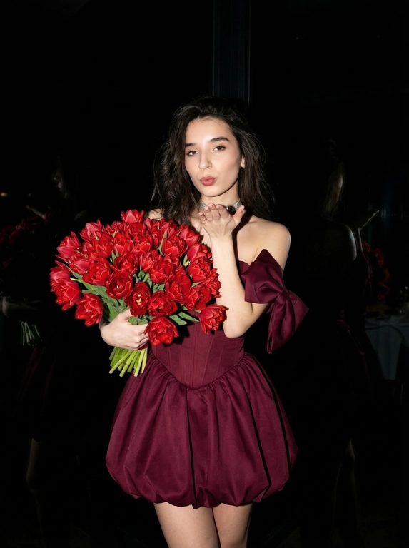 Сохранить внешность на 100%. Черты лица не изменять.
Ultra-realistic vertical 9:16 photo, Y2K luxury nightlife, paparazzi flash.

Полная темнота.
Фон почти полностью чёрный — ресторан не читается, только намёки: слабые отражения, едва заметные силуэты, глянец поверхностей. Всё пространство «съедено» тьмой.

Единственный источник света — жёсткая вспышка камеры, бьёт прямо в лицо и фигуру, создаёт резкий контраст, глубокие тени, эффект ночного paparazzi-кадра начала 2000-х.

Женщина в платье:
— глубокий винно-бордовый / dark cherry цвет
— корсетное мини без бретелей
— жёсткий sculptural corset
— короткая объёмная bubble-юбка
— off-shoulder банты
— плотная дорогая couture-ткань
— силуэт 1:1, без изменений

В руках — огромный, сверхпышный букет цветов, почти перекрывает корпус:
— тяжёлый, статусный,
— максимально плотный,
— luxury florist look.

Действие:
— она посылает воздушный поцелуй в камеру,
— взгляд дерзкий, холодный, уверенный,
— без улыбки.

Эффект:
— вспышка «вырывает» её из темноты,
— кожа и ткань ловят свет,
— фон проваливается в чёрный.

Стиль:
Y2K rich bitch, flash-only lighting, dark luxury, paparazzi moment, ultra-realistic, expensive, iconic, no softness, no romance, pure power.

Какие будут цветы: Пионовидные тюльпаны красного цвета, большой букет. Девушка держит его обеими руками. Волосы распущены.