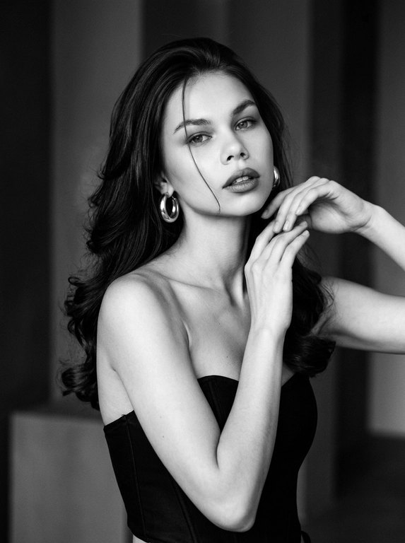 Кинематографический эффект с добавлением зерна. 
Вертикальный fashion-портрет, чёрно-белая плёночная съёмка в стиле editorial. 
Кадр по пояс, лёгкий поворот корпуса, плечи по диагонали, взгляд в камеру. Техника - 85mm, Kodak Tri-X 400, мягкая тональность, сильное зерно, легкий шум и немного дыма. Драматичные тени. Безупречный макияж, губы с более тёмным контуром, слегка приоткрыты. Лёгкое движение головы и корпуса с деликатным motion blur,  подбородок приподнят, руки у лица.
В ушах толстые серьги кольца.
Чёрный корсетный топ без бретелей, акцент на плечах и шее. Мягкий студийный свет спереди и сбоку, плавные тени. Ультрареализм, натуральная текстура кожи, monochrome film editorial aesthetic.

Какая будет укладка? : Волосы каскадом, брашинг с объёмом у корней, концы подкручены внутрь, одна прядь падает на лицо.

Доп. характеристики: длинные черные волосы