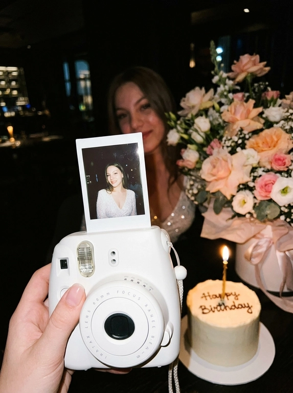 Edit the uploaded reference image into a photorealistic, luxury
birthday party scene.
Preserve the person’s identity exactly. Keep their real facial
structure, skin tone, age presentation, hair type, body type, natural
proportions, expression style, and distinctive features. The final
image should still clearly look like the same person from the
reference photo.
Create the image in the style of a dark, intimate iPhone UGC birthday
snapshot taken in a dim room with direct front flash. Use a dynamic
handheld camera angle, slightly tilted or off-center, as if the photo
was taken spontaneously at the party. The perspective should feel
natural, candid, and immersive, with the white instant camera with
polaroid angled and in focus of the generated image. Avoid a stiff,
perfectly centered studio composition.
The mood should feel candid, and special, like a real birthday
moment captured casually at night. Use flash-lit foreground details,
darker background shadows, warm candle glow, subtle phone-
camera grain, slight handheld imperfection, and shallow depth of
field.
The main focus must be a freshly printed instant polaroid photo
coming out of a clean white instant camera in the foreground. The
white camera should be large in the lower foreground, held close to
the lens. The polaroid should be sharp, visually dominant, and
positioned with a natural slight angle for a more dynamic
composition. The composition should feel similar to a casual iPhone
photo where the polaroid and camera are the main subject, while the
actual person behind it is only subtly visible, and not the main focus,
face not visible.
A natural, well-groomed hand matching the person’s skin tone and
overall styling holds the white instant camera. The hand should look
realistic and relaxed, with simple polished nails only if suitable for
the person.
Inside the polaroid, show the same person from the reference image
posing elegantly at their birthday celebration. They should look
confident, refined, and warmly expressive. Transform their outfit into
a polished birthday look that suits their appearance and gender
presentation. For a woman, create an elegant dress or refined
celebratory outfit. For a man, create a fashionable formal or semi-
formal outfit suitable for a luxury birthday setting. The outfit color should
harmonize with or be inspired by the outfit color in the original
reference photo.
Behind the polaroid, show only a subtle blurred glimpse of the same
person in the background, wearing the same transformed outfit. The
background person should be softly out of focus, partly hidden by
the polaroid camera.
Include tasteful birthday décor around the scene. Use elegant
flowers that harmonize with the subject’s outfit and the overall color
palette of the reference photo. The flowers may be roses, orchids,
hydrangeas, baby’s breath, peonies, lilies, greenery, or other refined
florals. Choose colors that match the person’s transformed outfit
and create a cohesive luxury birthday atmosphere.
Place a birthday cake to the side or slightly in the background. The
cake should be visible but secondary to the polaroid. The cake color,
icing, floral decorations, and candle color should harmonize with the
person’s outfit and the overall palette. Add one lit candle. The cake
must clearly say exactly: “Happy Birthday” in neat, readable icing.
Use realistic direct flash highlights, intimate dark-room lighting, warm
candlelight, soft party lights, shallow depth of field, realistic skin
texture, natural facial proportions, elegant styling, and a cohesive
color palette based on the reference image.
**Strict constraints:** no identity change, no altered facial structure,
no mismatched person between the polaroid and background, no
extra people, no rings, no bracelets, no hand jewelry, no distorted
hands, no extra fingers, no duplicated faces, no warped camera, no
unreadable cake writing, no misspelled cake text, no extra text, no
overly bright studio lighting, no flat commercial lighting, no plastic
skin, no stiff straight-on composition

Доп. характеристики: Без макияжа точь в точь как на фотографии