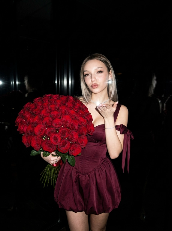 Сохранить внешность на 100%. Черты лица не изменять.
Ultra-realistic vertical 9:16 photo, Y2K luxury nightlife, paparazzi flash.

Полная темнота.
Фон почти полностью чёрный — ресторан не читается, только намёки: слабые отражения, едва заметные силуэты, глянец поверхностей. Всё пространство «съедено» тьмой.

Единственный источник света — жёсткая вспышка камеры, бьёт прямо в лицо и фигуру, создаёт резкий контраст, глубокие тени, эффект ночного paparazzi-кадра начала 2000-х.

Женщина в платье:
— глубокий винно-бордовый / dark cherry цвет
— корсетное мини без бретелей
— жёсткий sculptural corset
— короткая объёмная bubble-юбка
— off-shoulder банты
— плотная дорогая couture-ткань
— силуэт 1:1, без изменений

В руках — огромный, сверхпышный букет цветов, почти перекрывает корпус:
— тяжёлый, статусный,
— максимально плотный,
— luxury florist look.

Действие:
— она посылает воздушный поцелуй в камеру,
— взгляд дерзкий, холодный, уверенный,
— без улыбки.

Эффект:
— вспышка «вырывает» её из темноты,
— кожа и ткань ловят свет,
— фон проваливается в чёрный.

Стиль:
Y2K rich bitch, flash-only lighting, dark luxury, paparazzi moment, ultra-realistic, expensive, iconic, no softness, no romance, pure power.

Какие будут цветы: В руках у нее букет тёмно-красных роз.