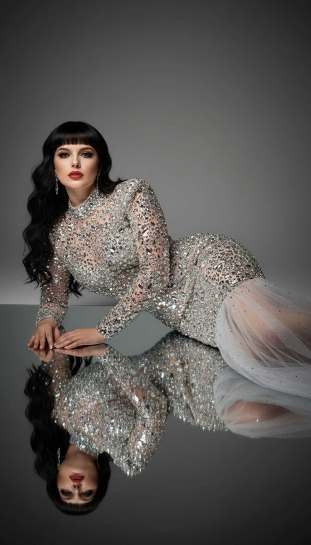 Это изысканный портрет женщины, лежащей боком на зеркальной поверхности, создавая идеальное отражение всего ее тела. На ней элегантное, облегающее платье от кутюр, полностью расшитое серебряными стразами, кристаллами и сверкающими деталями, которые роскошно ловят свет. Платье имеет длинные рукава, высокий ворот и струящийся шлейф из прозрачного тюля, усыпанного стразами на юбке, источающий гламур и изысканность. У нее длинные, волнистые волосы, которые нежно падают на ее плечи. Ее макияж безупречен с яркими темными smoky eyes и черной подводкой для глаз, длинными, объемными ресницами, безупречной кожей и пухлыми губами с изысканной, яркой красной помадой. На ней сдержанные, но элегантные серьги, дополняющие гламурный образ. Сцена драматически освещена темной виньеткой, подчеркивающей блеск платья и контраст между ее телом и отражением в зеркале. Минималистичный фон в нейтральных тонах ещё больше подчёркивает харизму модели и богатство деталей наряда. Общая атмосфера создаёт ощущение роскоши, элегантности и изысканности, отражая моду и высокую моду. Сохранены те же черты лица без каких-либо изменений, тот же тон кожи и цвет волос. Качественный маникюр, профессиональное студийное качество изображения.