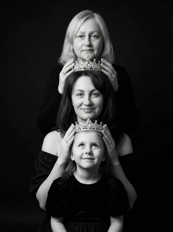 Это студийный портрет, выполненный в драматичном черно-белом стиле. Фон черный. В кадре три женщины разного возраста: две женщины и один ребёнок. Они расположены вертикально друг над другом, образуя «башню». Каждая женщина одета в темную одежду, у всех одежда разного стиля. Верхняя женщина в топе с длинными рукавами, у нее корона на голове . Следующая в платье с открытыми плечами. И ребёнок в платье с рукавами-фонариками.
У каждой на голове корона, которую предыдущая женщина держит руками. Все смотрят в камеру с нежным выражением лица, с лёгкой улыбкой Освещение мягкое, акцент сделан на лицах. Цветовая гамма монохромная, с преобладанием черного и серебристого (от корон). Атмосфера торжественная и немного загадочная. Не меняй черты лица, цвет волос и цвет глаз.

