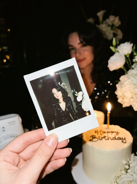 Edit the uploaded reference image into a photorealistic, luxury
birthday party scene.
Preserve the person’s identity exactly. Keep their real facial
structure, skin tone, age presentation, hair type, body type, natural
proportions, expression style, and distinctive features. The final
image should still clearly look like the same person from the
reference photo.
Create the image in the style of a dark, intimate iPhone UGC birthday
snapshot taken in a dim room with direct front flash. Use a dynamic
handheld camera angle, slightly tilted or off-center, as if the photo
was taken spontaneously at the party. The perspective should feel
natural, candid, and immersive, with the white instant camera with
polaroid angled and in focus of the generated image. Avoid a stiff,
perfectly centered studio composition.
The mood should feel candid, and special, like a real birthday
moment captured casually at night. Use flash-lit foreground details,
darker background shadows, warm candle glow, subtle phone-
camera grain, slight handheld imperfection, and shallow depth of
field.
The main focus must be a freshly printed instant polaroid photo
coming out of a clean white instant camera in the foreground. The
white camera should be large in the lower foreground, held close to
the lens. The polaroid should be sharp, visually dominant, and
positioned with a natural slight angle for a more dynamic
composition. The composition should feel similar to a casual iPhone
photo where the polaroid and camera are the main subject, while the
actual person behind it is only subtly visible, and not the main focus,
face not visible.
A natural, well-groomed hand matching the person’s skin tone and
overall styling holds the white instant camera. The hand should look
realistic and relaxed, with simple polished nails only if suitable for
the person.
Inside the polaroid, show the same person from the reference image
posing elegantly at their birthday celebration. They should look
confident, refined, and warmly expressive. Transform their outfit into
a polished birthday look that suits their appearance and gender
presentation. For a woman, create an elegant dress or refined
celebratory outfit. For a man, create a fashionable formal or semi-
formal outfit suitable for a luxury birthday setting. The outfit color should
harmonize with or be inspired by the outfit color in the original
reference photo.
Behind the polaroid, show only a subtle blurred glimpse of the same
person in the background, wearing the same transformed outfit. The
background person should be softly out of focus, partly hidden by
the polaroid camera.
Include tasteful birthday décor around the scene. Use elegant
flowers that harmonize with the subject’s outfit and the overall color
palette of the reference photo. The flowers may be roses, orchids,
hydrangeas, baby’s breath, peonies, lilies, greenery, or other refined
florals. Choose colors that match the person’s transformed outfit
and create a cohesive luxury birthday atmosphere.
Place a birthday cake to the side or slightly in the background. The
cake should be visible but secondary to the polaroid. The cake color,
icing, floral decorations, and candle color should harmonize with the
person’s outfit and the overall palette. Add one lit candle. The cake
must clearly say exactly: “Happy Birthday” in neat, readable icing.
Use realistic direct flash highlights, intimate dark-room lighting, warm
candlelight, soft party lights, shallow depth of field, realistic skin
texture, natural facial proportions, elegant styling, and a cohesive
color palette based on the reference image.
**Strict constraints:** no identity change, no altered facial structure,
no mismatched person between the polaroid and background, no
extra people, no rings, no bracelets, no hand jewelry, no distorted
hands, no extra fingers, no duplicated faces, no warped camera, no
unreadable cake writing, no misspelled cake text, no extra text, no
overly bright studio lighting, no flat commercial lighting, no plastic
skin, no stiff straight-on composition