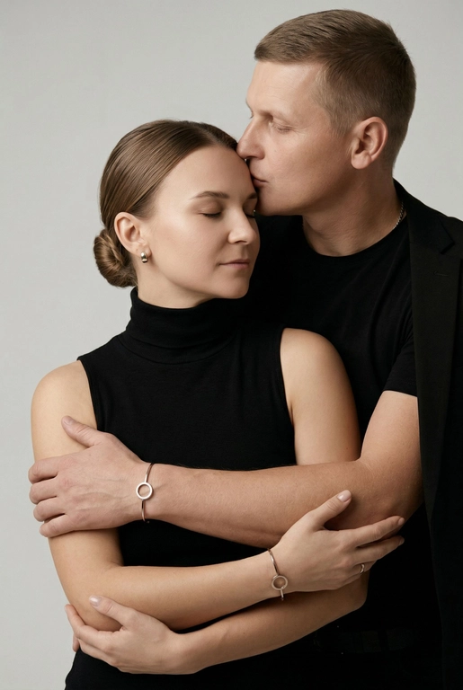 Мужчина и женщина позируют в студии в объятиях. Мужчина стоит позади женщины, обнимая её обеими руками; его тело повернуто к ней, голова слегка наклонена вниз, губы касаются её лба. Женщина немного отклоняет голову назад к его плечу, её глаза закрыты. Кадр — средний, от талии до головы, снят на уровне глаз. Композиция плотная, акцентирует линии рук и соприкосновение лиц. Женщина стоит, прямо обращённая к камере.
На ней облегающий чёрный топ без рукавов с высоким воротом, из плотной матовой ткани.
Мужчина одет в чёрную футболку и чёрную неброскую куртку. На обоих запястьях — тонкие металлические браслеты с минималистичными круглыми подвесками, а женщина носит небольшие светлые металлические серьги с гладкой поверхностью.
Обстановка минималистичная: однотонный светло-серый фон без текстуры.
Освещение — мягкий студийный свет: рассеянный фронтальный и боковой свет создаёт плавные переходы света и тени на лицах и руках. Тени мягкие, средней глубины. Контраст умеренный, без жёстких бликов. Свет равномерно распределён по фигурам, фон подсвечен мягко, без выраженных градиентов.
Съёмка на портретный объектив 85 мм, малая глубина резкости с чётким фокусом на лицах и руках; фон слегка размыт, текстуры кожи и ткани высоко детализированы. Это минималистичный студийный портрет, высокодетализированный и фотореалистичный.
Минималистичное романтическое студийное настроение, нейтральные тёплые тона кожи, низкая–средняя насыщенность, мягкое рассеянное освещение, средний контраст с нежными тенями, плавные переходы бликов, естественная передача кожи, чистый светлый фон, тонкий тональный баланс, современная мягкая редакционная цветокоррекция.

Волосы женщины собраны в низкий, гладкий пучок с аккуратно уложенными прядями.
