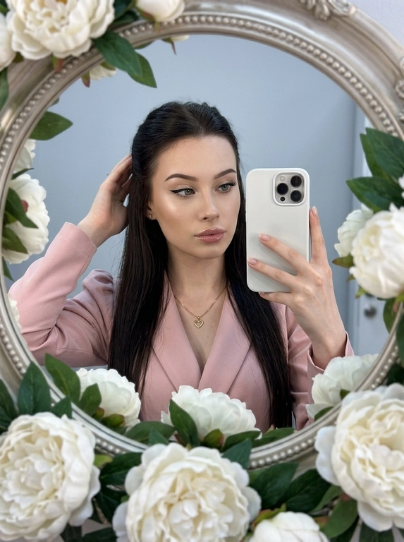 Максимально реалистичное селфи в зеркале в эстетике luxury soft glam. Модель стоит перед круглым зеркалом с изящной рамой, украшенной белыми живыми пионами. Цветы обрамляют зеркало нежно и дорого, создавая ощущение утончённого, премиального кадра. Композиция чистая и визуально собранная: модель находится в центре отражения, кадр выглядит эстетично, дорого и современно.

Поза естественная и уверенная: одной рукой модель держит современный смартфон, второй рукой слегка касается волос, создавая живой и непринуждённый жест. Селфи выглядит не постановочным, а как красивый спонтанный кадр из дорогой life-style съёмки.

Макияж — выразительный soft glam: чёткие чёрные стрелки, длинные пышные ресницы с лисьим эффектом, внутренние уголки глаз подсвечены сияющими тенями. Кожа ровная, ухоженная, с мягким влажным сиянием. Губы подчёркнуты карандашом и покрыты полупрозрачным глянцевым блеском, создающим аккуратный объём.

На модели ее образ выглядит женственно, современно и дорого. 

Смартфон современный, в минималистичном белом матовом чехле. Если нужен более дорогой вайб, без лишнего декора и ярких деталей.

Освещение холодное, мягкое и чистое, как в дорогой beauty-съёмке или premium dressing room. Свет красиво подчеркивает сияние кожи, текстуру ткани, свежесть пионов и блеск украшения. Атмосфера — роскошная, женственная, ухоженная, без дешёвого глянца.

Фотореализм максимальный: высокая детализация кожи, ткани, цветов, аксессуаров и отражения в зеркале. Лёгкий цифровой шум, мягкая резкость, деликатная зернистость, естественная глубина кадра, высокая контрастность, эффект живого фото, без CGI и без искусственного AI-эффекта.

Какая будет прическ?: полусобранная укладка, верхняя часть волос аккуратно убрана назад, мягкие пряди обрамляют лицо.

Какой будет образ на фото? : Элегантный пудрово-розовый жакет с акцентными рукавами-фонариками и глубоким V-образным вырезом. На шее — тонкая изящная цепочка с ювелирным кулоном в форме сердца.