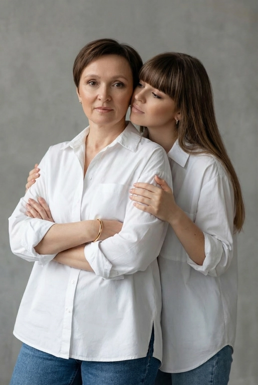 Используй эти два фото мамы и дочери и создай для них студийный портрет в чистом и элегантном стиле. Они стоят рядом, очень близко друг к другу. Дочь мягко обнимает маму за руку и слегка к ней прижимается со спины. Мама сложила руки перед грудью. Образ минималистичный, современный и стильный. На руке у матери тонкий золотой браслет. Профессиональный макияж и прическа.  Не менять черты лица ни на 1 процент. Фон — нейтральный серый с мягкой текстурой. Освещение рассеянное, мягкое, подчёркивающее естественные тона кожи. Атмосфера тёплая, нежная и спокойная, в стиле журнальной съёмки. Обе в свободных стильных белых рубашках с подвернутыми рукавами и джинсах.