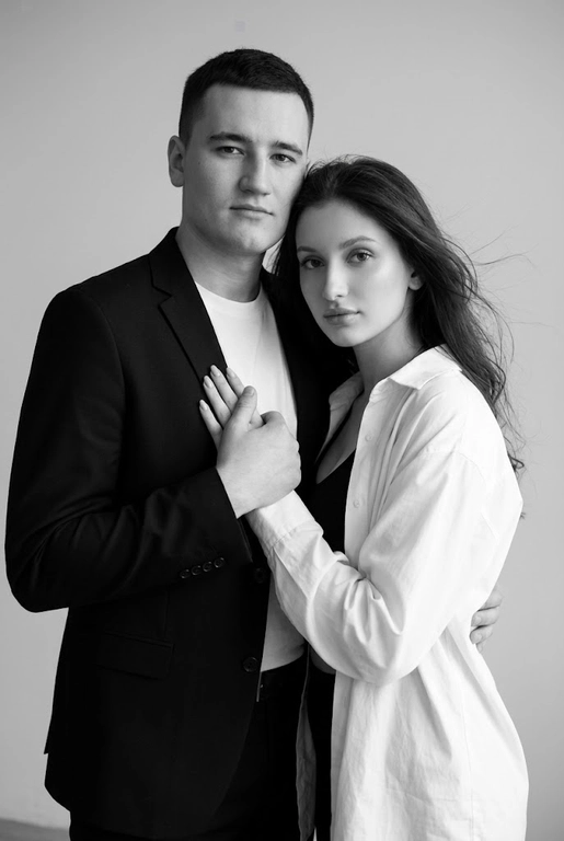 Черно-белый портрет мужчины и женщины в стиле фэшн фотосессии в фотостудии. Не изменяйте лица. Фон нейтральный, однотонный, светло-серый. Атмосфера интимная и спокойная. На переднем плане женщина. Вся композиция выполнена в приглушенной монохромной гамме со студийным освещением, которое подчеркивает текстуру кожи и одежды. Её руки лежат на груди мужчины. Мужчина обнимает женщину сзади, положив свою руку поверх её руки.

Она одета в черный топ, белую оверсайз рубашку. Мужчина одет в  классическом черном пиджаке и белой футболке.