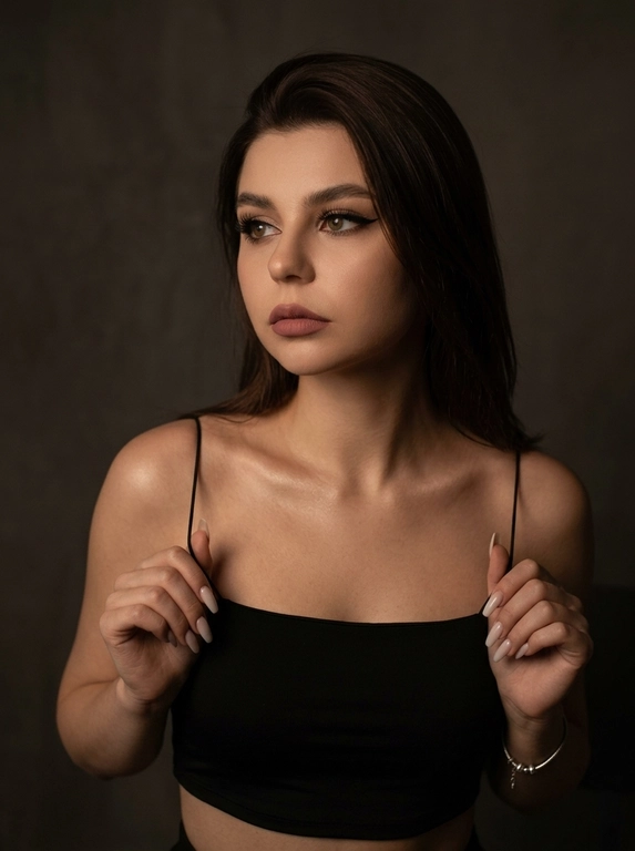 Максимально реалистичная фотография в стиле moody boudoir editorial, передающая атмосферу интимности, чувственности и приглушенного драматизма.
Крупный план по пояс (waist-up shot / close-up). женщина позирует в полумраке, придерживая руками края одежды. [Поза/взгляд/эмоция]: Плечи слегка приподняты, руки с длинным светлым маникюром сжимают тонкие бретели черного топа; голова немного повернута в сторону, взгляд задумчивый и отведен в сторону; выражение лица томное, загадочное и уверенное.
Вечерний «glam» выразительные стрелки, густо накрашенные ресницы, создающие эффект «кошачьего взгляда», мягкий контуринг лица и матовая помада цвета пыльной розы.
Облегающий черный кроп-топ на очень тонких бретелях-спагетти. Материал — матовая эластичная ткань. На запястье виден изящный серебряный браслет.
Минималистичный интерьер в темных тонах. Фон однотонный, слегка текстурированный, уходящий в глубокую тень, что создает эффект камерности.
 Низкий ключ (low-key lighting). Мягкий, теплый направленный свет падает сбоку, подчеркивая рельеф ключиц, линию плеч и текстуру кожи. Глубокие, кинематографичные тени создают объем и драматичное настроение.
 85mm f/1.4, идеальное фокусное расстояние для портрета, создающее минимальные искажения и сильное размытие фона. Очень малая глубина резкости; фокус идеально наведен на глаза и лицо модели, в то время как плечи и руки уходят в мягкое расфокусирование.
Гиперреалистичная текстура кожи: видны поры, естественный блеск на выступающих частях лица и плеч. Отсутствие «пластикового» эффекта ретуши.
 текстура ткани топа под пальцами, тонкие волоски на макушке, мягкий градиент тени на стене.
Эстетика «dark feminine energy», интимная, теплая и дорогая атмосфера (expensive moody vibe).

Какая будет прическа?: Волосы: перекинутые на одно плечо. Текстура волос гладкая и шелковистая, с отдельными тонкими прядями, подсвеченными теплым светом.

Доп. характеристики: не меняй мои черты лица