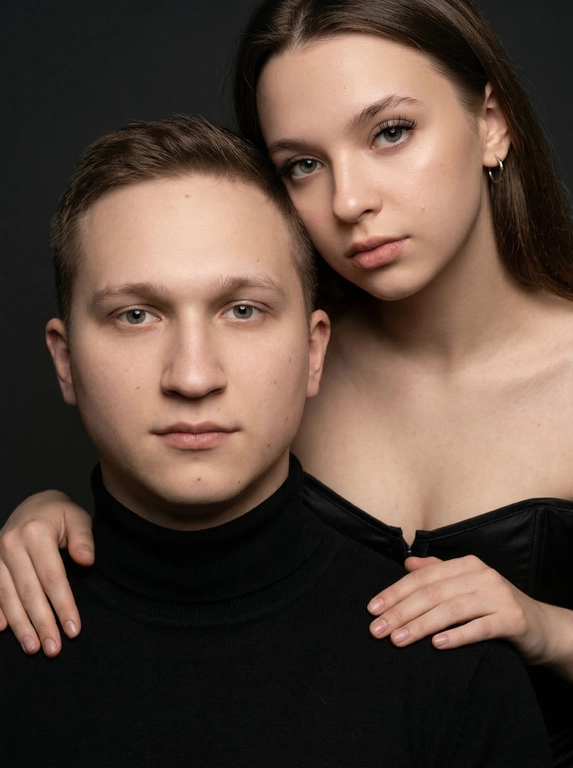 Гиперреалистичный студийный fashion-портрет молодой пары по загруженному референсу, 100% сходство лиц. Черты лица не изменять. Крупный план, кадр по плечи. Мужчина, спокойный уверенный взгляд прямо в камеру, естественное выражение лица, одет в черный трикотажный свитер с высоким воротом. Женщина стоит позади мужчины и нежно обнимает его, ее руки лежат на его плечах. Ее лицо расположено рядом с его лицом, она повернута слегка боком, голова немного наклонена вперед, взгляд направлен прямо в камеру. У женщины выразительные глаза с длинными густыми черными ресницами, естественный clean-glam макияж, натуральные брови, теплый оливковый оттенок кожи, небольшие серьги-кольца. Композиция плотная и интимная, их лица расположены очень близко друг к другу, современный модный портрет пары. Женщина одета в черный корсет. Кожа максимально натуральная: живая текстура кожи, видимые микропоры, subtle skin glow, естественный мягкий блеск кожи, natural skin microtexture, skin translucency, micro skin details, легкие естественные несовершенства кожи, без ретуши и без эффекта пластика. Освещение: мягкий студийный beauty lighting, рассеянный ключевой свет спереди и немного сбоку, мягкие глубокие тени для объема лица, деликатные световые блики на коже. Фон: глубокий темный студийный фон, темно-графитовый или почти черный, создающий контраст с кожей и лицами. Стиль: luxury fashion editorial photography, high fashion portrait, ultra photorealistic, 85mm portrait lens, shallow depth of field, creamy bokeh, cinematic soft lighting, RAW photo, extremely detailed, fine hair strands, natural skin pores, professional photography, photorealism, 8k, high dynamic range.
