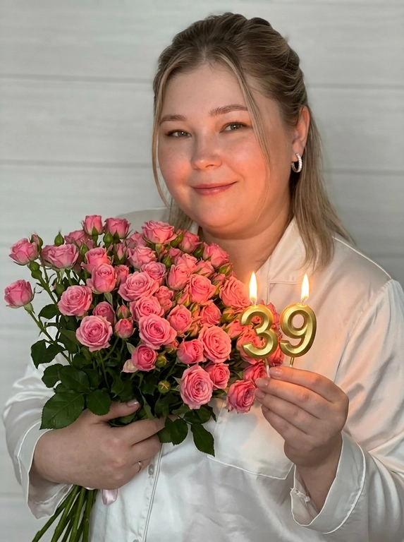 Вертикальный экстремально крупный цветной портрет по пояс в мягком романтичном стиле, камера на уровне груди, лёгкий фронтальный ракурс со смещением вправо, малая глубина резкости, фокус на руках и золотых зажжённых свечах-цифрах, образующих число .
Модель стоит полубоком, корпус в три четверти, плечи расслаблены, взгляд в камеру, милая полуулыбка, нежный наклон к цветам. В ближней к камере руке она держит свечи, вытянув руку вперёд в нижнюю центральную часть кадра, пальцы аккуратно обнимают ножки свечей, пламя даёт тёплый локальный свет. Вторая рука прижимает к груди плотный объёмный очень большой букет мелких ярко-розовых кустовых роз, занимающий левую половину кадра, почти без видимой зелени.  маленькие широкие бриллиантовые серьги-кольца.
Белое платье-кимоно с опущенными плечами и длинными широкими рукавами / расслабленная белая шёлковая пижама. Фон - светлая стена с легкой горизонтальной текстурой, немного размытая. Мягкий тёплый свет, как от ламп или бокового софтбокса, лёгкий акцент на свечах и руках, жёлто-оранжевые отблески, мягкие тени, уютная мечтательная атмосфера нежного birthday-портрета.

Какое будет число на свечах?: 39

Какая будет прическа?: Причёска: верхняя часть волос убрана назад, остальные распущены, слегка растрёпанные, прямой пробор, по две тонкие пряди у лица,
