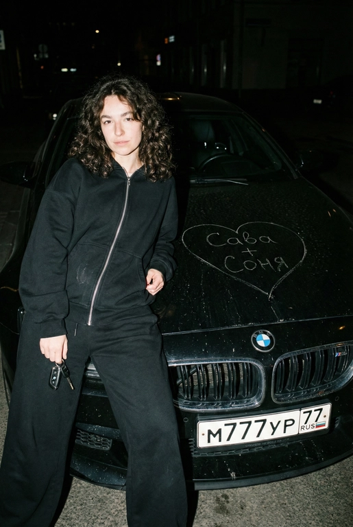 Строго сохранить лицо с прикреплённой референс-фотографии, на 100% без искажений. Человек одет полностью в чёрный, монохромный оверсайз спортивный костюм: свободная худи на молнии и широкие расслабленные штаны, без логотипов, текста и брендинга. Акцент на реальной текстуре кожи и детализированной текстуре ткани.
Сырой кинематографичный ночной кадр. Человек опирается на капот автомобиля, средний план, лёгкий верхний ракурс. В одной руке игриво держит ключи от машины. Номерной знак виден: m777yp 77.
Строго по центру капота, покрывая всю поверхность капота, глубоко и грубо процарапаны (не мелом) царапины: большое сердце с текстом «» внутри.
Жёсткая прямая вспышка, сильные тени, высокий контраст, видимая пыль и микро-царапины на краске. Тёмный размытый городской фон.
Объектив 35mm, f/2.8, грубая уличная эстетика.

Надпись на капоте: Сава + Соня

Марка автомобиля: Опирается на чёрный BMW M6 F90