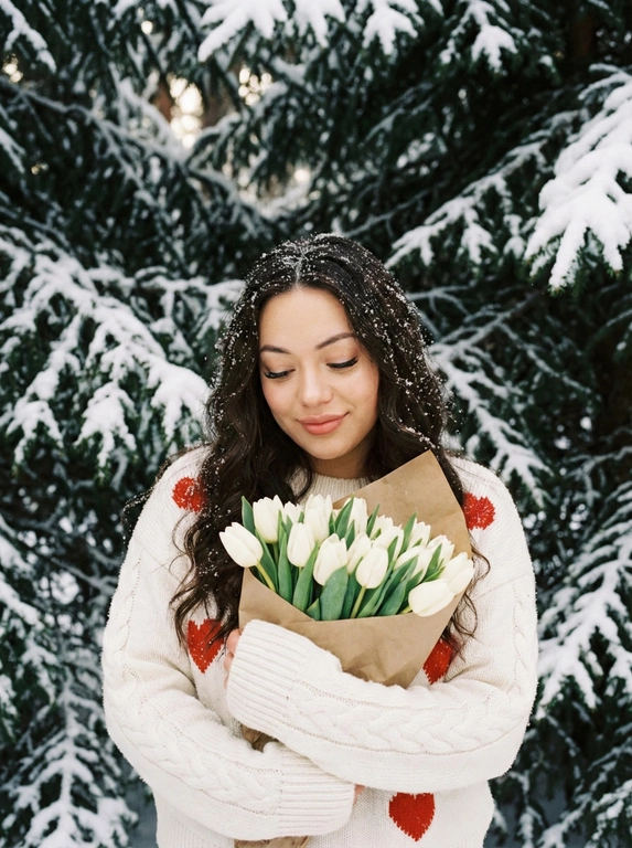   Привлекательная зимняя портретная фотография в стиле модной 35-миллиметровой пленки, сохраняйте свою внешность, девушка с длинными волнистыми волосами смотрит в камеру, глаза опущенны вниз на букет, милой легкой улыбкой, розовыми губами. Белый пушистый свитер с маленькими косами с красными сердечками, рукава падают на кисти. На руках - большой букет белых тюльпанов, завернутый в бумагу, прижатый к груди. Волосы и свитер, ферментированные свежим снегом. Фон - толстый, покрытый снегом, темно-зеленый, с объемной текстурой, толстые, плотно покрытые снегом, еловые ветви покрыты снегом, как в сказочном лесу. Прозрачный: мягкий диффузный дневной, теплый на коже, легкое свечение от снега, без теней. Цвета: чистый белый + ярко-красный +... Макияж в стиле мягкий глэм, сияющая кожа, акцент на большие...
