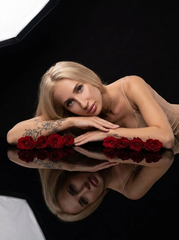 Студийный портрет женщины. Обстановка минималистичная: черный, глянцевый стол, отражающий человека и объекты на нем. Стиль фото - гламурная съемка с акцентом на красоту. Атмосфера спокойная и немного загадочная. Цветовая гамма - сочетание глубокого черного фона, красных роз и телесных оттенков кожи.

Женщина лежит на столе, опираясь на предплечья. Руки сложены, как будто она придерживает голову. На ней надет топ или платье с тонкими бретельками телесного цвета. На столе, вдоль рук человека, лежат красные розы. Они расположены таким образом, чтобы создавать симметричную композицию. Отражение в столе дублирует изображение человека и роз, усиливая эффект глубины и симметрии. Свет мягкий, рассеянный, акцентирует внимание на лице и розах, создавая игру света и теней.