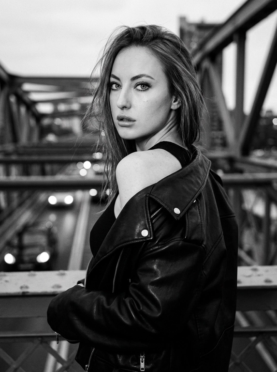 Используй внешность модели с фото. Фотореалистичное черно-белое фото в стиле модной редакционной съемки с эстетикой пленочной фотографии (зернистость, высокий контраст). Модель с волосами слегка растрепанными ветром. Она одета в черную, объемную кожаную куртку-косуху, которая небрежно спущена с левого плеча, открывая его. Под курткой надет черный облегающий топ. Поза: модель стоит вполоборота спиной к камере, но резко оборачивается и смотрит назад через плечо прямо в объектив. Выражение лица серьезное, дерзкое и чувственное, губы слегка приоткрыты, густые ресницы. Фон размыт, видны элементы конструкции моста и огни проезжающих машин вдали, создающие городскую атмосферу. Освещение естественное, подчеркивающее текстуру кожи и блеск кожи куртки. Высокое разрешение, 8k. Освещение мягкое и равномерное, без жестких теней, свет подчеркивает блеск жемчуга и страз, цвета насыщенные, но реалистичные. Высокая детализация, фотореализм, четкий фокус на лице, естественные пропорции.