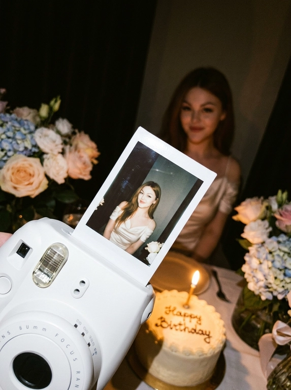 Edit the uploaded reference image into a photorealistic, luxury
birthday party scene.
Preserve the person’s identity exactly. Keep their real facial
structure, skin tone, age presentation, hair type, body type, natural
proportions, expression style, and distinctive features. The final
image should still clearly look like the same person from the
reference photo.
Create the image in the style of a dark, intimate iPhone UGC birthday
snapshot taken in a dim room with direct front flash. Use a dynamic
handheld camera angle, slightly tilted or off-center, as if the photo
was taken spontaneously at the party. The perspective should feel
natural, candid, and immersive, with the white instant camera with
polaroid angled and in focus of the generated image. Avoid a stiff,
perfectly centered studio composition.
The mood should feel candid, and special, like a real birthday
moment captured casually at night. Use flash-lit foreground details,
darker background shadows, warm candle glow, subtle phone-
camera grain, slight handheld imperfection, and shallow depth of
field.
The main focus must be a freshly printed instant polaroid photo
coming out of a clean white instant camera in the foreground. The
white camera should be large in the lower foreground, held close to
the lens. The polaroid should be sharp, visually dominant, and
positioned with a natural slight angle for a more dynamic
composition. The composition should feel similar to a casual iPhone
photo where the polaroid and camera are the main subject, while the
actual person behind it is only subtly visible, and not the main focus,
face not visible.
A natural, well-groomed hand matching the person’s skin tone and
overall styling holds the white instant camera. The hand should look
realistic and relaxed, with simple polished nails only if suitable for
the person.
Inside the polaroid, show the same person from the reference image
posing elegantly at their birthday celebration. They should look
confident, refined, and warmly expressive. Transform their outfit into
a polished birthday look that suits their appearance and gender
presentation. For a woman, create an elegant dress or refined
celebratory outfit. For a man, create a fashionable formal or semi-
formal outfit suitable for a luxury birthday setting. The outfit color should
harmonize with or be inspired by the outfit color in the original
reference photo.
Behind the polaroid, show only a subtle blurred glimpse of the same
person in the background, wearing the same transformed outfit. The
background person should be softly out of focus, partly hidden by
the polaroid camera.
Include tasteful birthday décor around the scene. Use elegant
flowers that harmonize with the subject’s outfit and the overall color
palette of the reference photo. The flowers may be roses, orchids,
hydrangeas, baby’s breath, peonies, lilies, greenery, or other refined
florals. Choose colors that match the person’s transformed outfit
and create a cohesive luxury birthday atmosphere.
Place a birthday cake to the side or slightly in the background. The
cake should be visible but secondary to the polaroid. The cake color,
icing, floral decorations, and candle color should harmonize with the
person’s outfit and the overall palette. Add one lit candle. The cake
must clearly say exactly: “Happy Birthday” in neat, readable icing.
Use realistic direct flash highlights, intimate dark-room lighting, warm
candlelight, soft party lights, shallow depth of field, realistic skin
texture, natural facial proportions, elegant styling, and a cohesive
color palette based on the reference image.
**Strict constraints:** no identity change, no altered facial structure,
no mismatched person between the polaroid and background, no
extra people, no rings, no bracelets, no hand jewelry, no distorted
hands, no extra fingers, no duplicated faces, no warped camera, no
unreadable cake writing, no misspelled cake text, no extra text, no
overly bright studio lighting, no flat commercial lighting, no plastic
skin, no stiff straight-on composition
