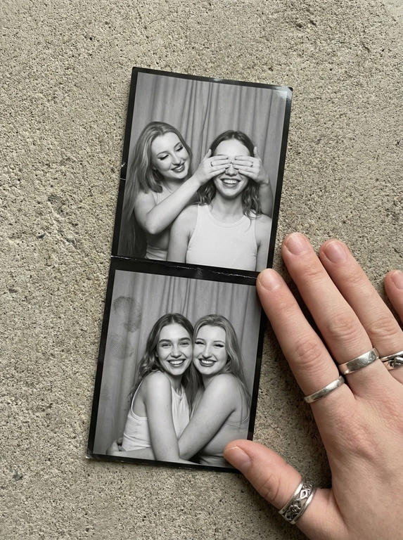   A vertical black-and-white photostrip lies casually on rough concrete near a hand adorned with silver rings, the contrast between the cool surface and monochrome tones enhancing authenticity. The strip showcases two women in simple tanks; in the first frame, one playfully covers the other's eyes, their smiles half-hidden and spontaneous. The following frame reveals both grinning fully into the lens, radiant and relaxed. Behind the subjects, a neutral curtain with faint folds and gentle texture creates the classic photobooth backdrop. The strip carries subtle creases and fingerprints, with soft shadows and muted highlights from a gentle iPhone flash. Slightly tilted and softly skewed, the composition feels like a genuine snapshot caught in an unguarded moment—contemporary photobooth snapshot, shot on iPhone.