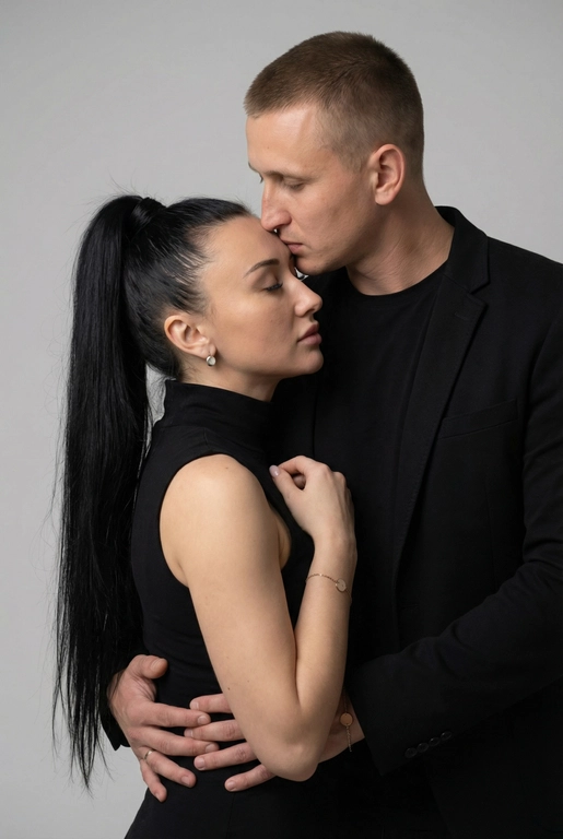   Мужчина и женщина позируют в студии в объятиях. Мужчина стоит позади женщины, обнимая её обеими руками; его тело повернуто к ней, голова слегка наклонена вниз, губы касаются её лба. Женщина немного отклоняет голову назад к его плечу, её глаза закрыты. Кадр — средний, от талии до головы, снят на уровне глаз. Композиция плотная, акцентирует линии рук и соприкосновение лиц. Женщина стоит, прямо обращённая к камере.
На ней облегающий чёрный топ без рукавов с высоким воротом, из плотной матовой ткани.
Мужчина одет в чёрную футболку и чёрную неброскую куртку. На обоих запястьях — тонкие металлические браслеты с минималистичными круглыми подвесками, а женщина носит небольшие светлые металлические серьги с гладкой поверхностью.
Обстановка минималистичная: однотонный светло-серый фон без текстуры.
Освещение — мягкий студийный свет: рассеянный фронтальный и боковой свет создаёт плавные переходы света и тени на лицах и руках. Тени мягкие, средней глубины. Контраст умеренный, без жёстких бликов. Свет равномерно распределён по фигурам, фон подсвечен мягко, без выраженных градиентов.
Съёмка на портретный объектив 85 мм, малая глубина резкости с чётким фокусом на лицах и руках; фон слегка размыт, текстуры кожи и ткани высоко детализированы. Это минималистичный студийный портрет, высокодетализированный и фотореалистичный.
Минималистичное романтическое студийное настроение, нейтральные тёплые тона кожи, низкая–средняя насыщенность, мягкое рассеянное освещение, средний контраст с нежными тенями, плавные переходы бликов, естественная передача кожи, чистый светлый фон, тонкий тональный баланс, современная мягкая редакционная цветокоррекция. Волосы женщины собраны в высокий хвост, длина до пояса 