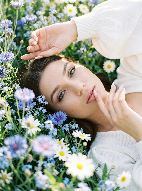 Экстремально крупный, кинематографичный портрет лица человека, лежащего среди густых полевых цветов, камера максимально близко — в кадре преимущественно лицо, черты сохранены без изменений, вокруг море цветов, создающее ощущение мягкого природного кокона, одна рука изящно поднята над головой, вторая мягко касается цветов рядом, на переднем плане сбоку частично размытые, не в фокусе цветы добавляют глубину и лёгкое ощущение движения, одета в летнее белое вязаное платье с объёмными рукавами, мягкая текстура ткани едва читается, естественный мягкий свет подчёркивает кожу и блики в волосах, малая глубина резкости, художественное боке, атмосферный кадр с деликатным плёночным зерном.



Какие будут цветы?: Цветы нежно-голубых, синих и белых оттенков.
