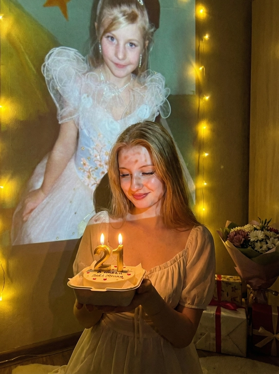       Создай ультрареалистичное фото, сделанное на камеру телефона в комнате, где изображен человек, который держит бенто торт с горящими золотыми свечами. В качестве фона на однотонную стену проецируется фотография из детства. Спроецированное фото занимает большую часть фона. Сочетание образов взрослого человека и ребёнка на фоне создаёт тёплую, ностальгическую атмосферу, подчеркивая связь между прошлым и настоящим. На стене висят гирлянды, рядом со стеной виднеются подарки и букет цветов. Снимок выглядит очень стильным и продуманным, присутствует ощущение праздника и воспоминаний. Не меняй внешность, полностью сохрани черты лица. Свечи на торте в форме числа 21. Женщина одета в молочное нежное платье. Черты лица не менять На фоне желтые новогодние гирлянды