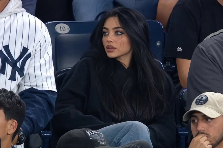 Person sitting in the crowd at a professional baseball game (or it could be any other match that I will write below), filmed from far away by a live broadcast camera. 
Seated in blue stadium chairs, slightly leaned back, looking off to the side with a surprised caught-on-camera expression, lips slightly parted.

Какая будет одежда? : Оверсайз худи черного цвета и джинсы
