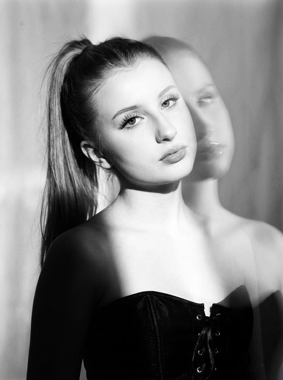 Вертикальный кадр. Художественный черно-белый портрет, снятый в светлой студии. Камера расположена на уровне лица, кадр крупный — портрет по плечи. Фон минималистичный, без деталей. Сохрани черты лица 1:1.  У модели естественный макияж, длинные натуральные ресницы, хайлайтер, аккуратный контуринг, глянцевый блеск на губах. На ней черный корсет без бретелей на завязках. Композиция — лицо слегка наклонено, взгляд направлен в камеру. Плечи и верхняя часть тела мягко уходят в тень, основной акцент на лице. Освещение — мягкий направленный солнечный естественный свет, падающий сбоку и сверху. Свет формирует плавный градиент между светом и глубокой тенью, подчёркивает структуру лица и создаёт кинематографичный контраст, солнечные блики на лице. Визуальный эффект — двойная экспозиция. Поверх основного чёткого портрета появляется полупрозрачный размытый силуэт головы той же модели, смещённый в сторону с эффектом моушн блюр. Этот второй слой выглядит как движение головы в момент съёмки — мягкий шлейф, создающий эффект динамики и призрачного дублирования лица. Текстура изображения — лёгкое плёночное зерно, мягкая дымка вокруг света, высокий контраст чёрно-белой фотографии. Атмосфера — минималистичный арт-портрет с экспериментальной эстетикой, ощущение движения и глубины. 


Какая будет прическа?: Волосы собраны в высокий хвост.