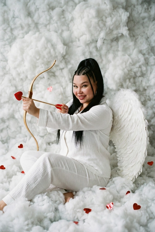 Использовать реальные черты лица человека с референса, естественную текстуру кожи и настоящий цвет и длину волос. Личность должна оставаться неизменной.

Y2K. Сказочная сцена с купидоном, сфотографированная как реальный момент.
Человек сидит среди плотных, пушистых белых облаков, которые полностью заполняют передний, средний и задний план, образуя бесшовную белую среду с многослойной глубиной.

Поза вполоборота: корпус слегка повернут вбок, одно плечо выдвинуто вперед, как будто человек натягивает и отпускает небольшой лук с красной стрелой в форме сердца; руки подняты с легким напряжением, указывающим на движение при выстреле.

Несмотря на поворот тела, голова повернута к объективу, взгляд направлен прямо в камеру. Выражение лица радостное и игривое, с мягкой улыбкой.

Наряд — простая белая пижама: свободная рубашка и брюки из муслина.
На спине — крылья из белых перьев, настоящие и фактурные, с четкими бликами по краям каждого пера.

Вокруг объекта в воздухе хаотично летают только конфетти в форме сердечек — маленькие красные сердечки из глянцевой фольги, расположенные на разном расстоянии: часть в переднем плане, часть растворяется среди облаков.

Без рассеянного света, без мягкого свечения, без студийного глоу.
Текстура кожи реалистичная и детализированная, без сглаживания и без фильтров.
Тонкая зернистость, волшебное, но искреннее ощущение Y2K-редакции — выглядит как фотография, а не визуализация.
