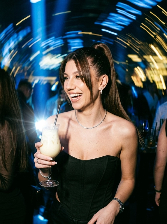 Фотореалистичный, хаотичный и откровенный кадр в стиле папарацци, снятый с прямой резкой вспышкой в переполненном элитном НОЧНОМ клубе. Великолепная роскошная женщина искренне смеется, держа в руке изящный бокал с коктейлем Пина Колада. На ней черный матовый корсет без бретелей и стильные серебряные аксессуары.
Главная особенность кадра: художественный эффект размытого движения (motion blur) на фоне. Ночные огни превращаются в живописные длинные световые треки и полосы насыщенного синего и теплого желтого цвета, создавая динамичный кадр света вокруг её четкой фигуры.
Объемный холодный свет. Текстура зернистой пленки (film grain), сырая энергия, атмосфера бурной ночной вечеринки, высокая детализация кожи и ткани.
-ar 4:5 -style raw


Какая будет прическа?: Гладкий высокий хвост и длинная челка-шторка, обрамляющая лицо.