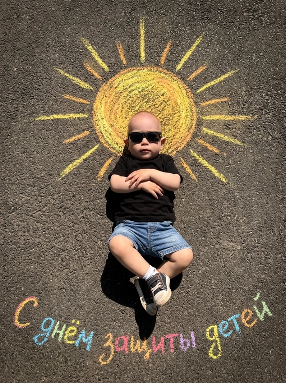 Top-down shot of a child lying on dark grainy asphalt. A bright chalk-drawn sun with radiant rays is drawn above the child’s head. The child is wearing black sunglasses, a black t-shirt, denim shorts, and sneakers with white socks. Arms crossed on chest, legs bent and crossed at ankles. Vibrant chalk sun texture in yellow, orange, and warm golden tones. Harsh midday sunlight, sharp shadows, high contrast. Summery playful atmosphere. On the asphalt below, colorful chalk lettering reads "С днем защиты детей". Photorealistic, 8k, highly detailed texture.