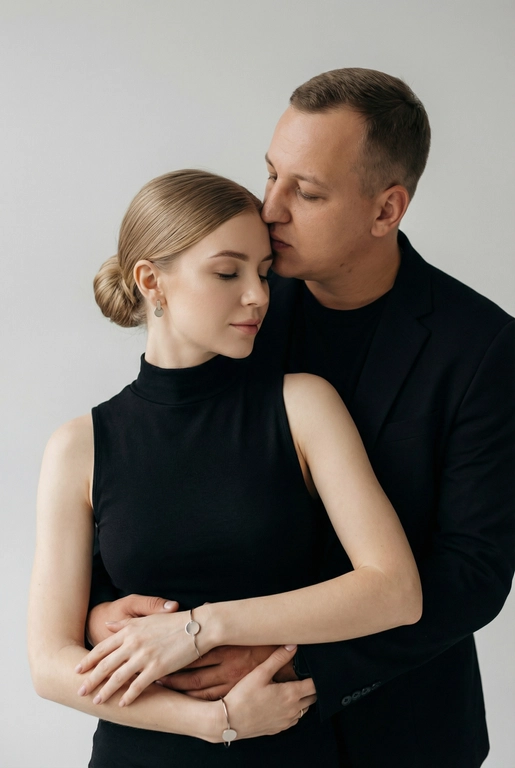   Мужчина и женщина позируют в студии в объятиях. Мужчина стоит позади женщины, обнимая её обеими руками; его тело повернуто к ней, голова слегка наклонена вниз, губы касаются её лба. Женщина немного отклоняет голову назад к его плечу, её глаза закрыты. Кадр — средний, от талии до головы, снят на уровне глаз. Композиция плотная, акцентирует линии рук и соприкосновение лиц. Женщина стоит, прямо обращённая к камере.
На ней облегающий чёрный топ без рукавов с высоким воротом, из плотной матовой ткани.
Мужчина одет в чёрную футболку и чёрную неброскую куртку. На обоих запястьях — тонкие металлические браслеты с минималистичными круглыми подвесками, а женщина носит небольшие светлые металлические серьги с гладкой поверхностью.
Обстановка минималистичная: однотонный светло-серый фон без текстуры.
Освещение — мягкий студийный свет: рассеянный фронтальный и боковой свет создаёт плавные переходы света и тени на лицах и руках. Тени мягкие, средней глубины. Контраст умеренный, без жёстких бликов. Свет равномерно распределён по фигурам, фон подсвечен мягко, без выраженных градиентов.
Съёмка на портретный объектив 85 мм, малая глубина резкости с чётким фокусом на лицах и руках; фон слегка размыт, текстуры кожи и ткани высоко детализированы. Это минималистичный студийный портрет, высокодетализированный и фотореалистичный.
Минималистичное романтическое студийное настроение, нейтральные тёплые тона кожи, низкая–средняя насыщенность, мягкое рассеянное освещение, средний контраст с нежными тенями, плавные переходы бликов, естественная передача кожи, чистый светлый фон, тонкий тональный баланс, современная мягкая редакционная цветокоррекция. Волосы женщины собраны в низкий, гладкий пучок с аккуратно уложенными прядями.