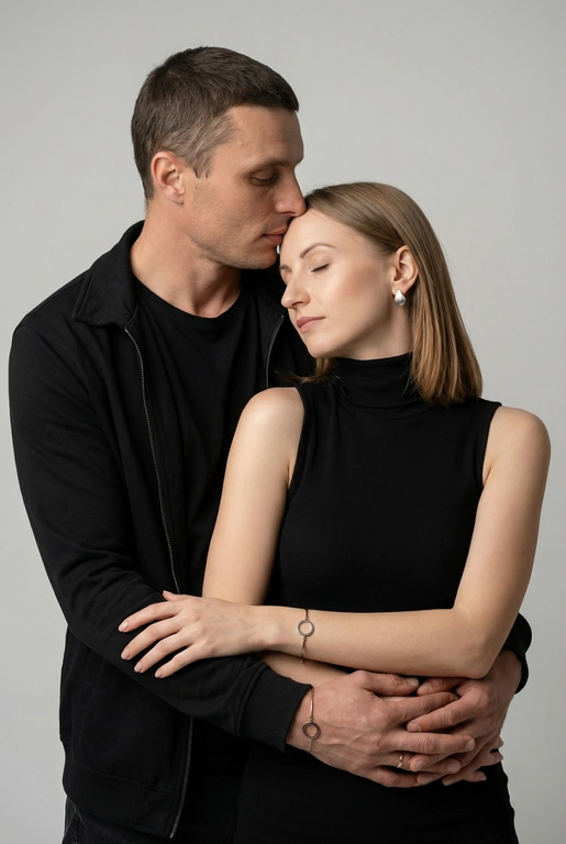   Мужчина и женщина позируют в студии в объятиях. Мужчина стоит позади женщины, обнимая её обеими руками; его тело повернуто к ней, голова слегка наклонена вниз, губы касаются её лба. Женщина немного отклоняет голову назад к его плечу, её глаза закрыты. Кадр — средний, от талии до головы, снят на уровне глаз. Композиция плотная, акцентирует линии рук и соприкосновение лиц. Женщина стоит, прямо обращённая к камере.
На ней облегающий чёрный топ без рукавов с высоким воротом, из плотной матовой ткани.
Мужчина одет в чёрную футболку и чёрную неброскую куртку. На обоих запястьях — тонкие металлические браслеты с минималистичными круглыми подвесками, а женщина носит небольшие светлые металлические серьги с гладкой поверхностью.
Обстановка минималистичная: однотонный светло-серый фон без текстуры.
Освещение — мягкий студийный свет: рассеянный фронтальный и боковой свет создаёт плавные переходы света и тени на лицах и руках. Тени мягкие, средней глубины. Контраст умеренный, без жёстких бликов. Свет равномерно распределён по фигурам, фон подсвечен мягко, без выраженных градиентов.
Съёмка на портретный объектив 85 мм, малая глубина резкости с чётким фокусом на лицах и руках; фон слегка размыт, текстуры кожи и ткани высоко детализированы. Это минималистичный студийный портрет, высокодетализированный и фотореалистичный.
Минималистичное романтическое студийное настроение, нейтральные тёплые тона кожи, низкая–средняя насыщенность, мягкое рассеянное освещение, средний контраст с нежными тенями, плавные переходы бликов, естественная передача кожи, чистый светлый фон, тонкий тональный баланс, современная мягкая редакционная цветокоррекция. Не изменять мою прическу