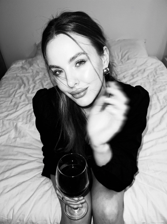 Create a portrait, transfer the face from the reference photo without changing facial features, proportions, or expression, preserve full identity. Aesthetic black-and-white nighttime Y2K photograph captured with direct on-camera flash, shot in motion.
A woman is sitting on the edge of a white, slightly rumpled bed, the frame taken from a slightly elevated angle. She wears a stylish black top with voluminous sleeves and large round statement earrings. In one hand she holds an elegant glass of dark wine. Her body is subtly moving, creating natural motion blur across the face and shoulders, as if the moment was caught mid-gesture. The gaze is languid and directed toward the camera, with a soft, intimate smile. Hair falls in large waves with loose strands drifting across the face, slightly displaced by movement. Lighting is harsh flash against deep night shadows, high contrast with blown highlights and strong falloff, visible film grain. Skin texture remains real and alive, no smoothing, enhanced by flash glare. The mood is high-fashion editorial, Parisian chic, old-money sensuality, nighttime melancholy, spontaneous and cinematic, shot in a raw 35mm film aesthetic with pronounced motion blur and lived-in realism.