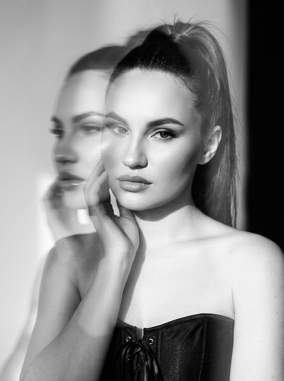 Вертикальный кадр. Художественный черно-белый портрет, снятый в светлой студии. Камера расположена на уровне лица, кадр крупный — портрет по плечи. Фон минималистичный, без деталей. Сохрани черты лица 1:1.  У модели естественный макияж, длинные натуральные ресницы, хайлайтер, аккуратный контуринг, глянцевый блеск на губах. На ней черный корсет без бретелей на завязках. Композиция — лицо слегка наклонено, взгляд направлен в камеру. Плечи и верхняя часть тела мягко уходят в тень, основной акцент на лице. Освещение — мягкий направленный солнечный естественный свет, падающий сбоку и сверху. Свет формирует плавный градиент между светом и глубокой тенью, подчёркивает структуру лица и создаёт кинематографичный контраст, солнечные блики на лице. Визуальный эффект — двойная экспозиция. Поверх основного чёткого портрета появляется полупрозрачный размытый силуэт головы той же модели, смещённый в сторону с эффектом моушн блюр. Этот второй слой выглядит как движение головы в момент съёмки — мягкий шлейф, создающий эффект динамики и призрачного дублирования лица. Текстура изображения — лёгкое плёночное зерно, мягкая дымка вокруг света, высокий контраст чёрно-белой фотографии. Атмосфера — минималистичный арт-портрет с экспериментальной эстетикой, ощущение движения и глубины. 


Какая будет прическа?: Волосы собраны в высокий хвост.