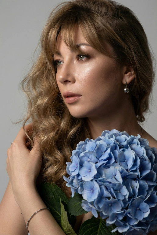 Создайте ультрареалистичный портрет, не меняя черты лица, крупным планом 3/4, с мягкой, освещённой кожей, атласной отделкой и естественным блеском, где женщина изображена в профиле. Спокойное и чувственное выражение лица, губы слегка полуоткрыты. Она носит серебряные серьги-шары и простой серебряный браслет на запястье.
Фон нейтральный, в светло-сером тоне, с тёмным, целенаправленным низким угловым освещением, которое подчёркивает контур лица, текстуру кожи и объём волос. Стиль редактирования — высокая мода, чёткий фокус 8K, реалистичная текстура, изысканная и элегантная атмосфера.


Длинные волнистые волосы покрывают часть лица объёмными локонами, выпадающими на лицо и плечо. 

Цветы: Держит в руках огромный букет голубых гортензий