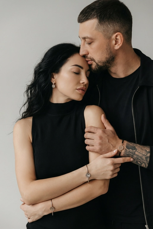   Мужчина и женщина позируют в студии в объятиях. Мужчина стоит позади женщины, обнимая её обеими руками; его тело повернуто к ней, голова слегка наклонена вниз, губы касаются её лба. Женщина немного отклоняет голову назад к его плечу, её глаза закрыты. Кадр — средний, от талии до головы, снят на уровне глаз. Композиция плотная, акцентирует линии рук и соприкосновение лиц. Женщина стоит, прямо обращённая к камере.
На ней облегающий чёрный топ без рукавов с высоким воротом, из плотной матовой ткани.
Мужчина одет в чёрную футболку и чёрную неброскую куртку. На обоих запястьях — тонкие металлические браслеты с минималистичными круглыми подвесками, а женщина носит небольшие светлые металлические серьги с гладкой поверхностью.
Обстановка минималистичная: однотонный светло-серый фон без текстуры.
Освещение — мягкий студийный свет: рассеянный фронтальный и боковой свет создаёт плавные переходы света и тени на лицах и руках. Тени мягкие, средней глубины. Контраст умеренный, без жёстких бликов. Свет равномерно распределён по фигурам, фон подсвечен мягко, без выраженных градиентов.
Съёмка на портретный объектив 85 мм, малая глубина резкости с чётким фокусом на лицах и руках; фон слегка размыт, текстуры кожи и ткани высоко детализированы. Это минималистичный студийный портрет, высокодетализированный и фотореалистичный.
Минималистичное романтическое студийное настроение, нейтральные тёплые тона кожи, низкая–средняя насыщенность, мягкое рассеянное освещение, средний контраст с нежными тенями, плавные переходы бликов, естественная передача кожи, чистый светлый фон, тонкий тональный баланс, современная мягкая редакционная цветокоррекция. Волосы женщины распучены и накручены в небрежную волну. 